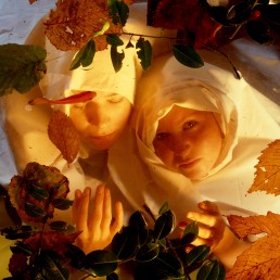 Two white woman, early 20s, cloaked in white, under water with leaves on top. Shot from above